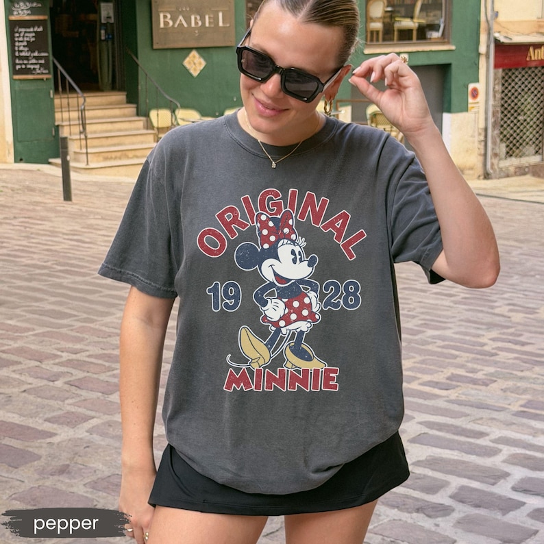 May include: Dark gray t-shirt featuring a Minnie Mouse graphic. The graphic includes the words "ORIGINAL MINNIE 1928" in red and blue. The shirt is a classic crew neck style.