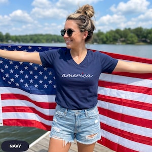 May include: Navy blue V-neck t-shirt with the word "america" in white script and a red star. The person is holding an American flag. The shirt is paired with denim shorts. The background includes a lake and trees.