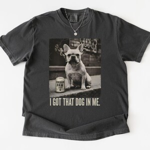 K&ouml;nnte beinhalten: Dunkelgraues T-Shirt mit einem Sepia-Bild einer franz&ouml;sischen Bulldogge mit Zigarette und einer Dose Urban Paw Lager. Der Text "I GOT THAT DOG IN ME." ist darunter gedruckt.