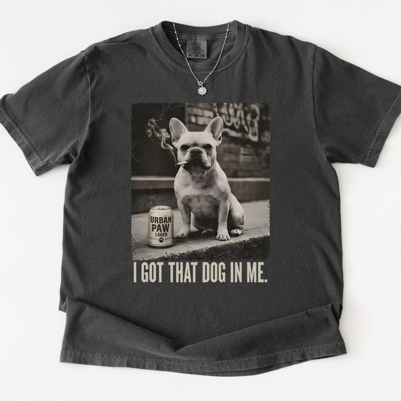 K&ouml;nnte beinhalten: Dunkelgraues T-Shirt mit einem Sepia-Bild einer franz&ouml;sischen Bulldogge mit Zigarette und einer Dose Urban Paw Lager. Der Text "I GOT THAT DOG IN ME." ist darunter gedruckt.