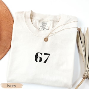 May include: Ivory-colored t-shirt with the number 67 printed in black. A gold necklace with a circular pendant rests on the shirt. The shirt is labeled "Comfort Colors". A brown hat and dried plant stems are in the background.