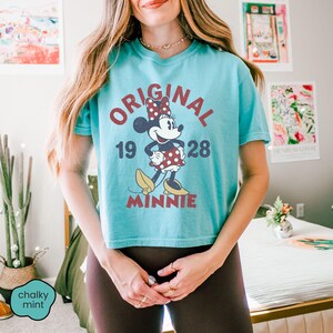 May include: A turquoise t-shirt featuring a graphic of Minnie Mouse with the text "ORIGINAL 1928 MINNIE". The design includes Minnie in a red polka dot dress and bow. The shirt is a cropped style.