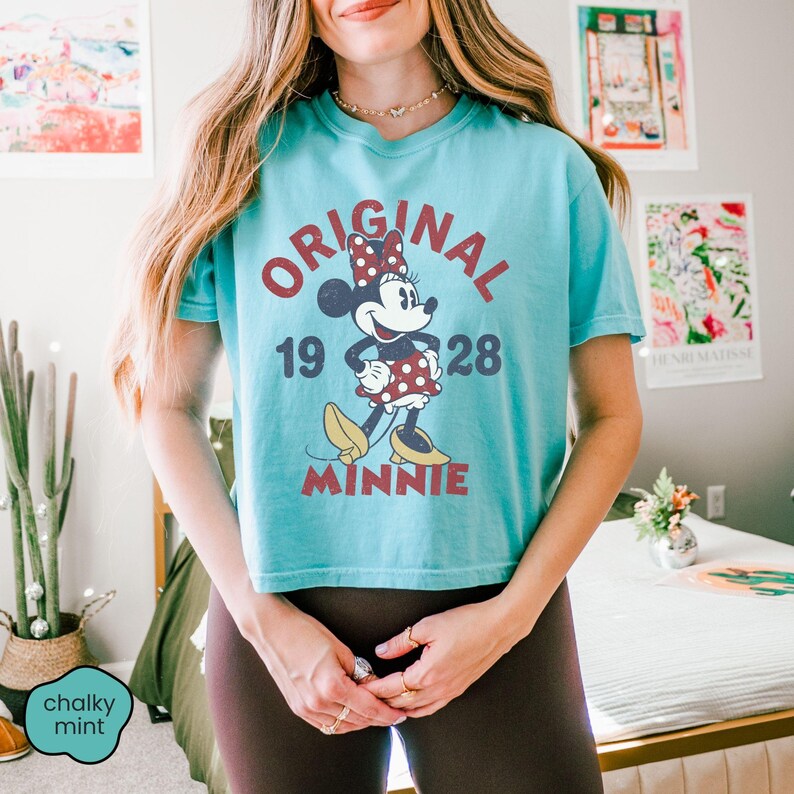 May include: A turquoise t-shirt featuring a graphic of Minnie Mouse with the text "ORIGINAL 1928 MINNIE". The design includes Minnie in a red polka dot dress and bow. The shirt is a cropped style.