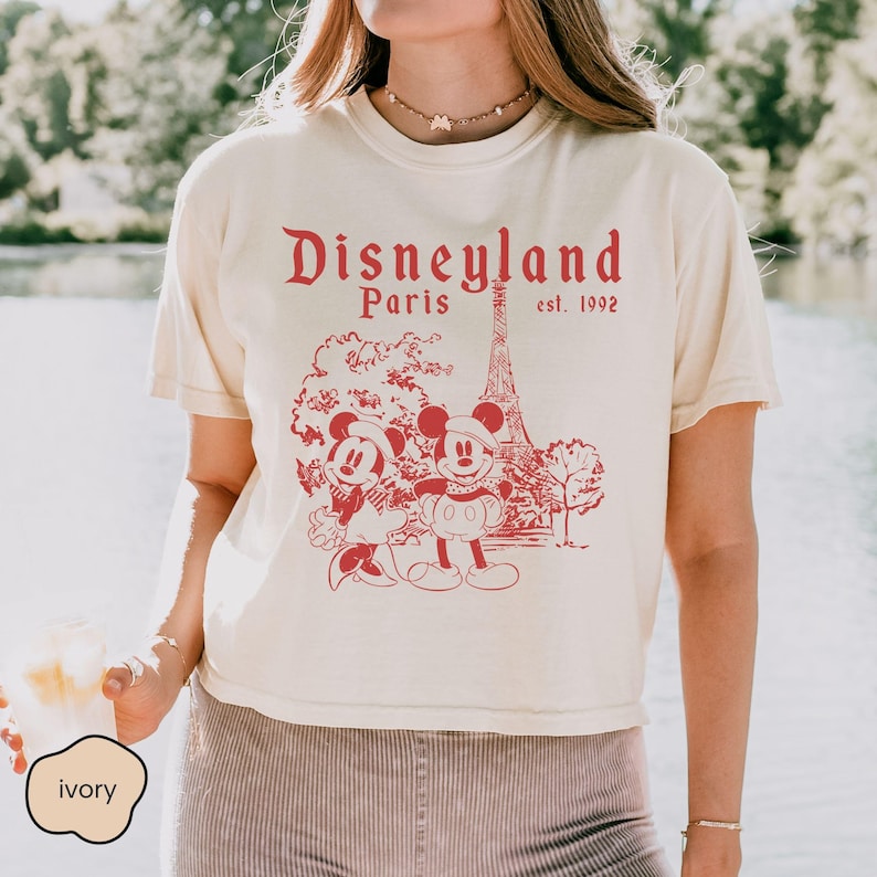 Puede incluir: Camiseta color marfil con un gr&aacute;fico rojo de Mickey y Minnie Mouse frente a la Torre Eiffel, con el texto "Disneyland Paris est. 1992". La camiseta tiene mangas cortas y cuello redondo.