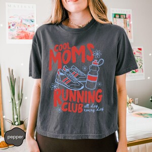 May include: A charcoal gray short-sleeve t-shirt featuring the red text COOL MOMS RUNNING CLUB and the blue text all day every day. The graphic includes a pair of running shoes and a water bottle, accented with small blue flower outlines.