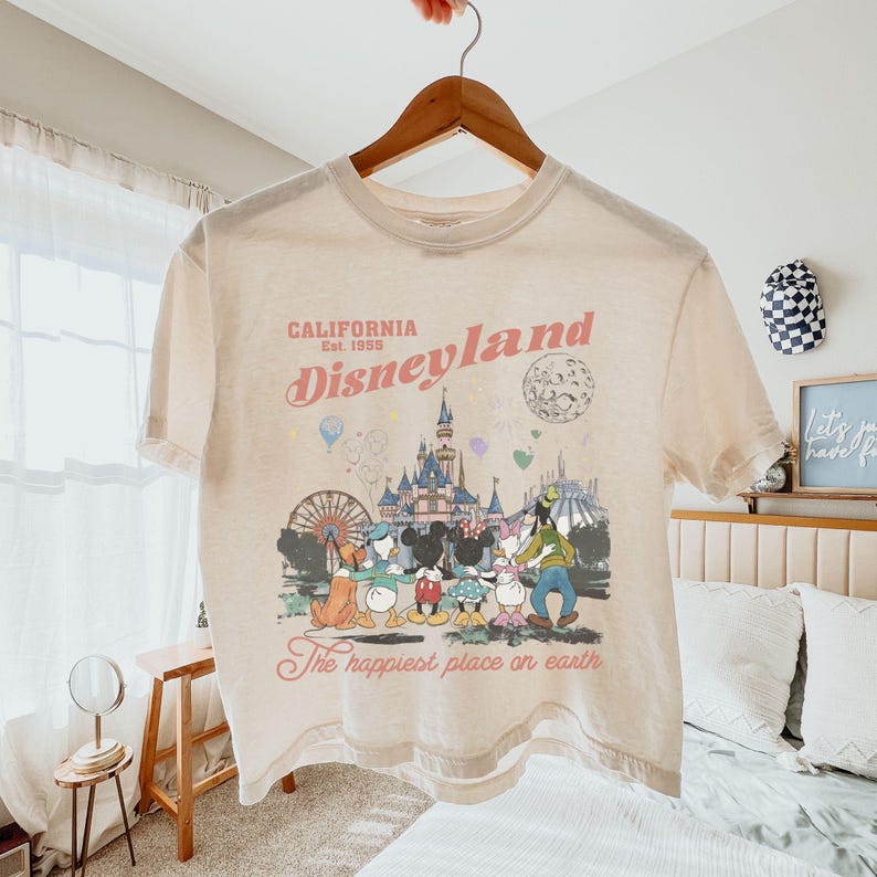 May include: Cream-colored t-shirt featuring a Disneyland graphic. The design includes the text "CALIFORNIA Est. 1955 Disneyland" above an illustration of Disney characters and a castle. The phrase "The happiest place on earth" is printed below the illustration.
