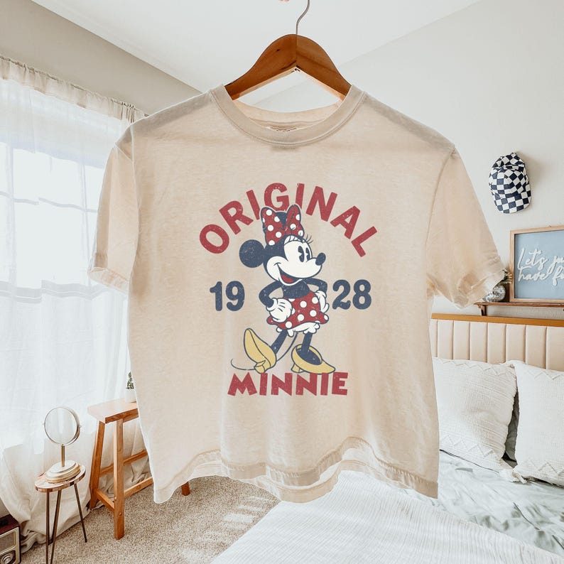 May include: Cream-colored t-shirt featuring a graphic of Minnie Mouse. The design includes the word "ORIGINAL" in red, the numbers "19 28", and the name "MINNIE". The shirt is hanging on a wooden hanger.