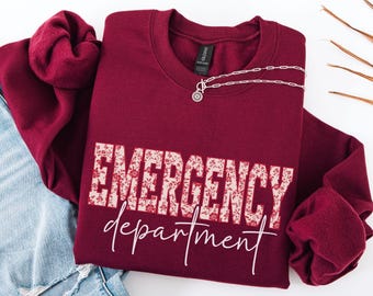 May include: A maroon sweatshirt with the words "EMERGENCY department" in a floral design. A silver necklace with a pendant is draped on the sweatshirt. The sweatshirt is folded on top of a pair of blue jeans.