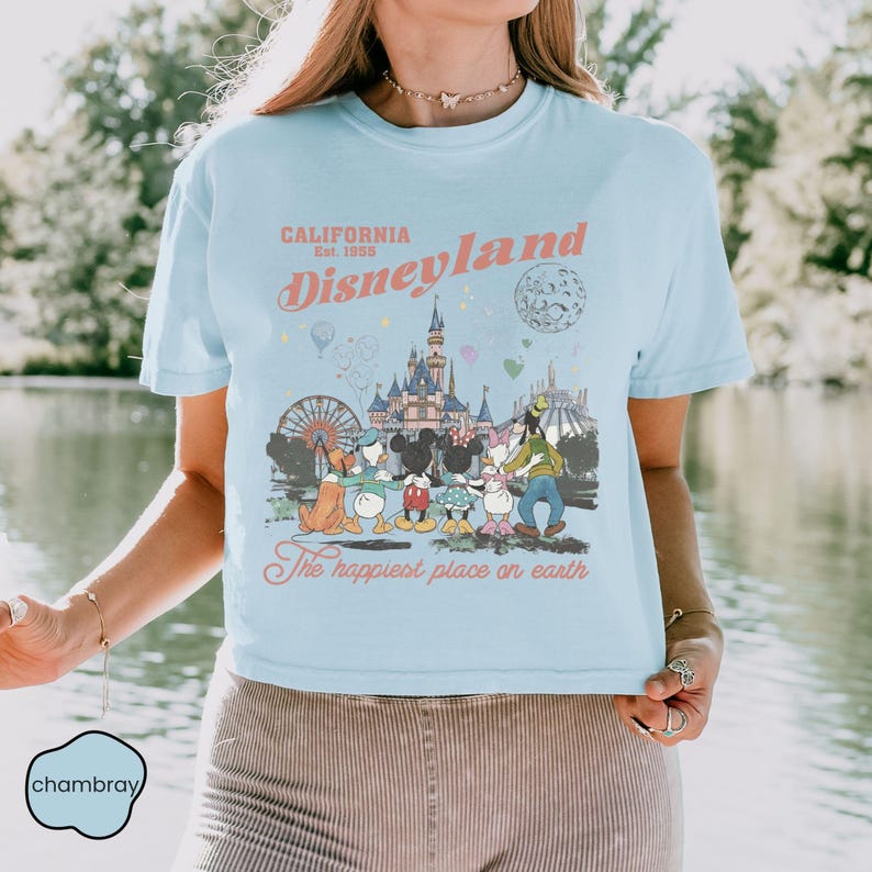 May include: Light blue t-shirt with a Disneyland graphic. The graphic features the text "California Disneyland Est. 1955" and an illustration of Disney characters in front of a castle and amusement park rides. The phrase "The happiest place on earth" is also included.