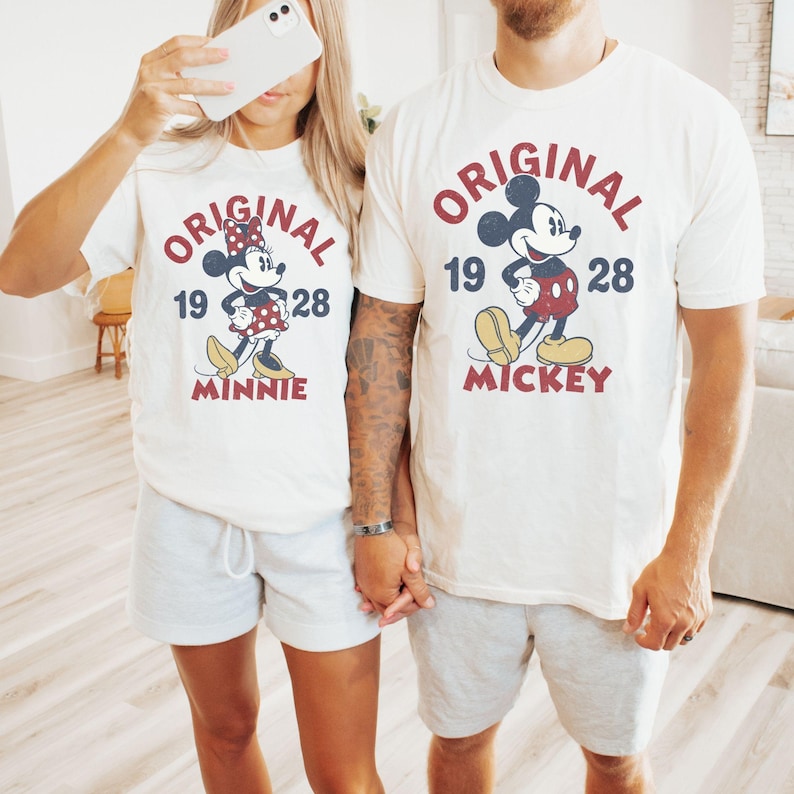 May include: Off-white t-shirts with matching gray shorts. Each shirt features a vintage-style graphic of Mickey and Minnie Mouse with the text "ORIGINAL 1928". The Mickey shirt says "MICKEY" and the Minnie shirt says "MINNIE".