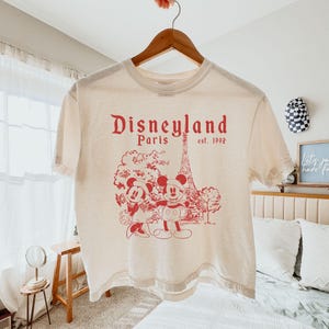 Puede incluir: Camiseta color crema con un gr&aacute;fico rojo de Mickey y Minnie Mouse frente a la Torre Eiffel, con el texto "Disneyland Paris est. 1992". La camiseta est&aacute; colgada en una percha de madera.