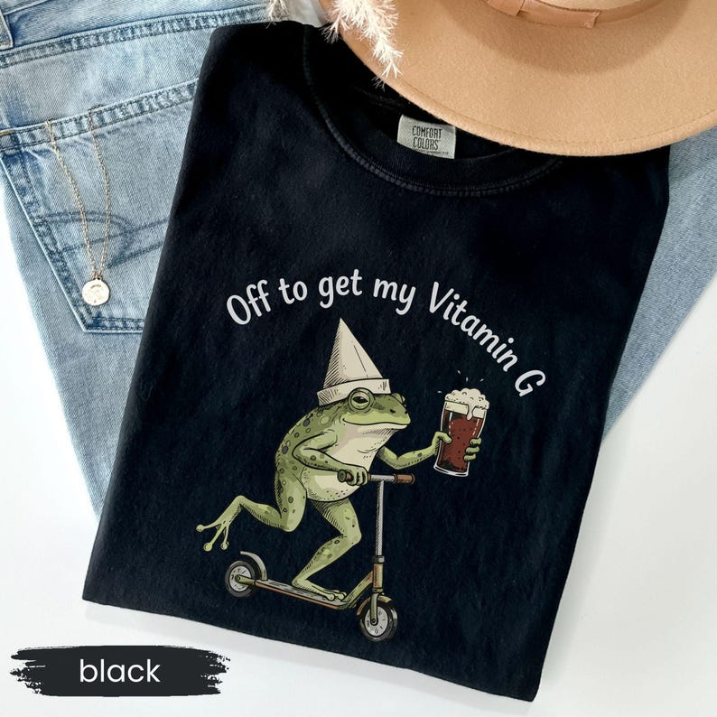 May include: Black t-shirt with the phrase "Off to get my Vitamin G." The graphic features a green frog wearing a hat and riding a scooter while holding a beer. The shirt is folded on top of blue jeans and a tan hat.