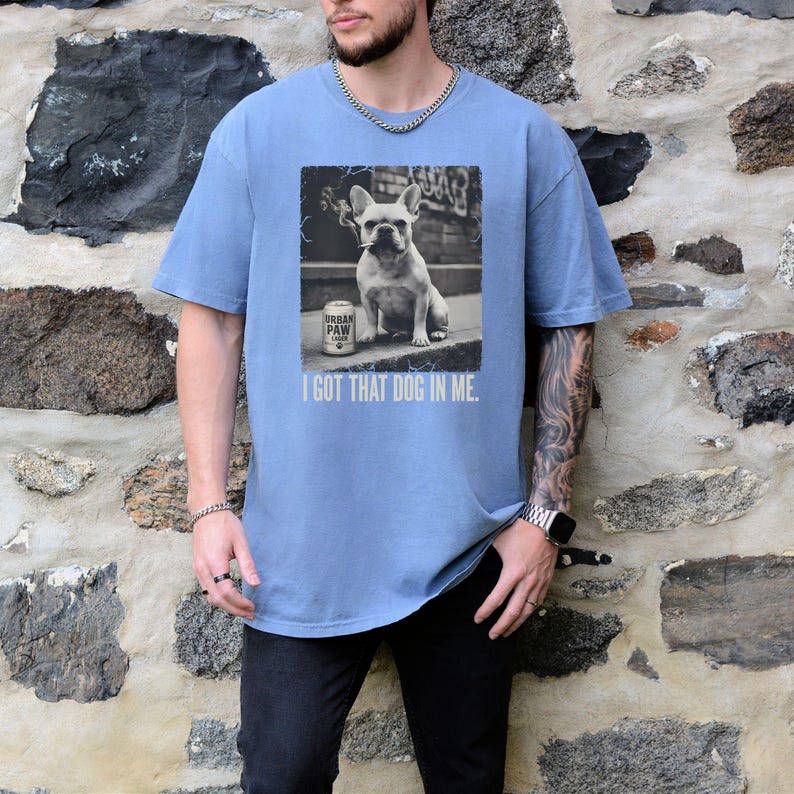 K&ouml;nnte beinhalten: Hellblaues T-Shirt mit einem Schwarz-Wei&szlig;-Bild einer franz&ouml;sischen Bulldogge mit einer Dose Urban Paw Bier. Der Text "I GOT THAT DOG IN ME" ist unter dem Bild aufgedruckt. Das Shirt ist aus weichem Material.