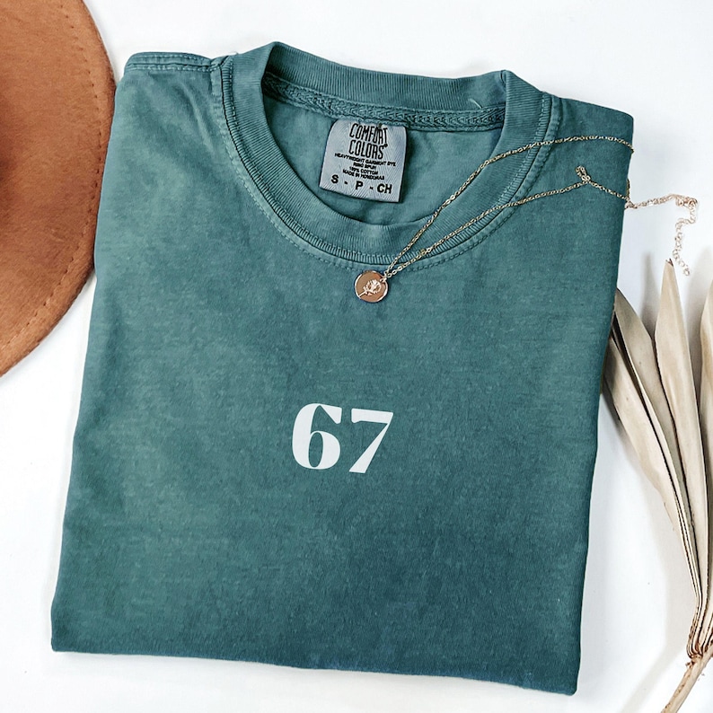 May include: A teal Comfort Colors t-shirt with the number 67 in white block letters. A gold necklace with a pendant rests on the shirt. A brown hat and dried plant are in the background.
