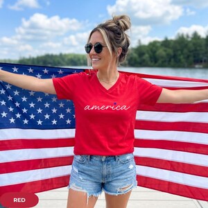 K&ouml;nnte beinhalten: Ein rotes V-Ausschnitt-T-Shirt mit dem Wort "america" in Schreibschrift und einem blauen Stern. Das Shirt wird mit Jeansshorts kombiniert. Im Hintergrund ist eine amerikanische Flagge zu sehen.