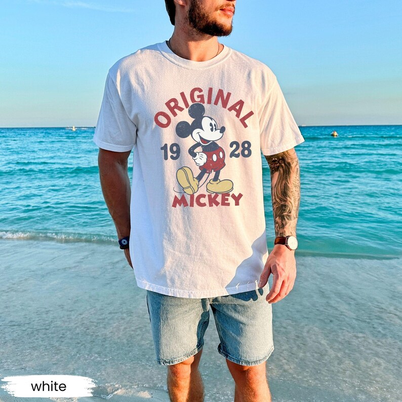 May include: White t-shirt featuring a vintage Mickey Mouse graphic. The design includes the word "ORIGINAL" in red, the numbers "19 28", and the name "MICKEY". The shirt is worn with denim shorts.