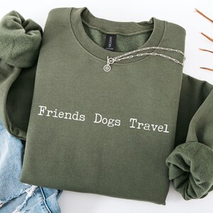 May include: An olive green sweatshirt with the words "Friends Dogs Travel" printed in white. A silver necklace with a small pendant is on the sweatshirt. The sweatshirt is folded on top of a pair of blue jeans.
