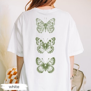 May include: White t-shirt featuring three green butterfly illustrations on the back. The butterflies are detailed with intricate wing patterns. The word "white" is printed at the bottom of the shirt.