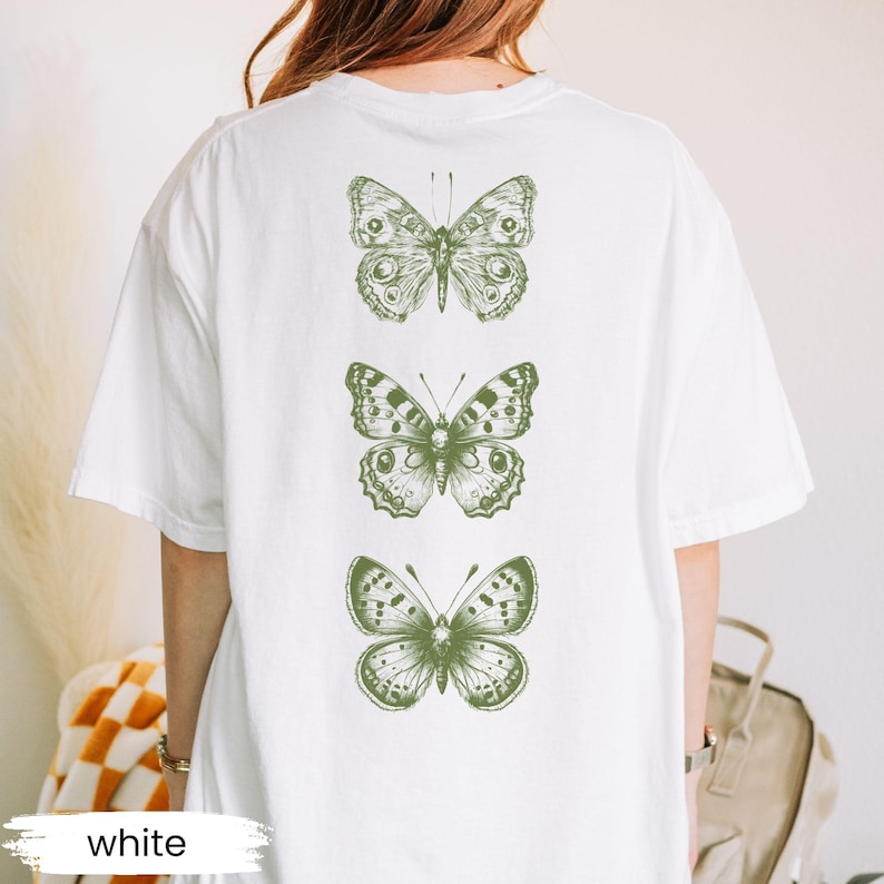 May include: White t-shirt featuring three green butterfly illustrations on the back. The butterflies are detailed with intricate wing patterns. The word "white" is printed at the bottom of the shirt.