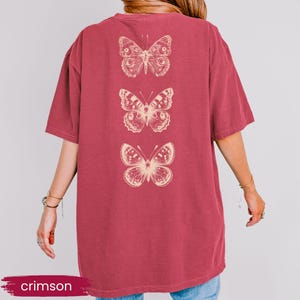 May include: Crimson-colored t-shirt featuring three detailed butterfly illustrations in a cream color. The butterflies are vertically aligned on the back of the shirt. The word "crimson" is displayed in a banner at the bottom.