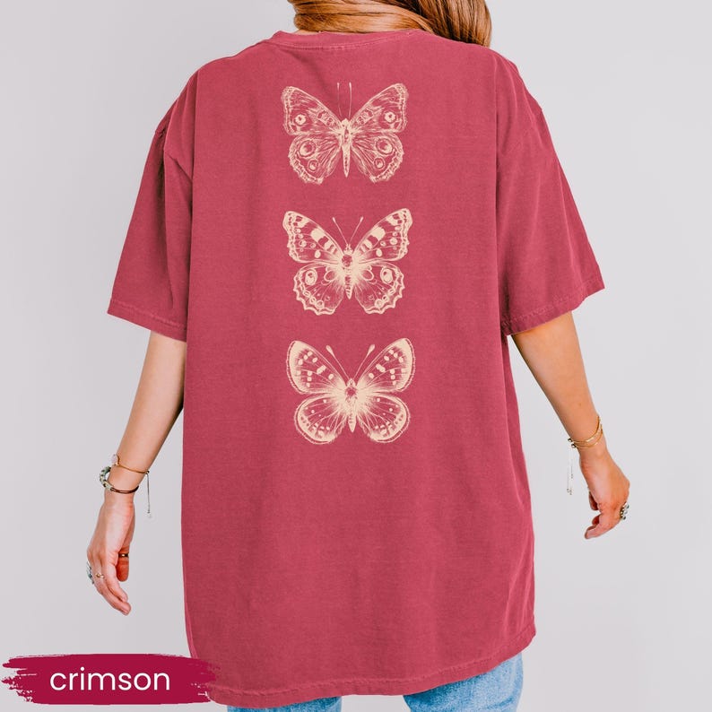 May include: Crimson-colored t-shirt featuring three detailed butterfly illustrations in a cream color. The butterflies are vertically aligned on the back of the shirt. The word "crimson" is displayed in a banner at the bottom.