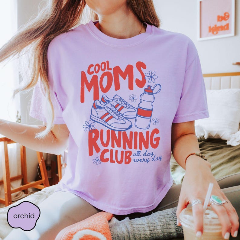 May include: A light purple t-shirt featuring a graphic print with the text COOL MOMS RUNNING CLUB all day every day, accompanied by illustrations of a pair of sneakers, a water bottle, and small flowers in red and blue ink colors.