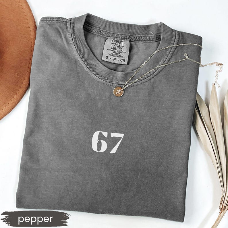 May include: A gray, short-sleeved t-shirt with the number 67 printed in white. The shirt has a crew neck and a small tag at the neckline. A silver necklace with a round pendant rests on the shirt. The word "pepper" is at the bottom.