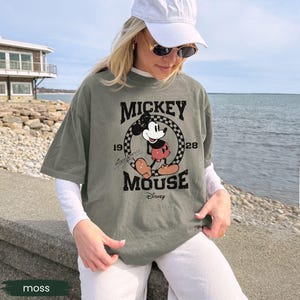 May include: Moss green t-shirt featuring a vintage Mickey Mouse graphic. The design includes the words "Mickey Mouse" and the year "1928" in a black, distressed font. The shirt is a relaxed fit and has short sleeves.
