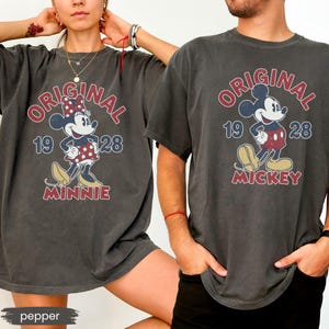 May include: Two charcoal gray t-shirts featuring vintage-style Mickey and Minnie Mouse designs. The shirts display the word "ORIGINAL" in red, the year "1928", and the character's name. The Minnie shirt has a red polka dot bow.