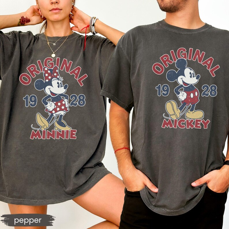 May include: Two charcoal gray t-shirts featuring vintage-style Mickey and Minnie Mouse designs. The shirts display the word "ORIGINAL" in red, the year "1928", and the character's name. The Minnie shirt has a red polka dot bow.