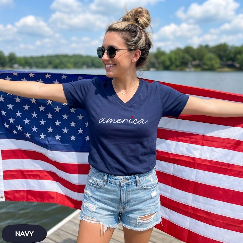 K&ouml;nnte beinhalten: Marineblaues V-Ausschnitt-T-Shirt mit dem Wort "america" in wei&szlig;er Schrift und einem roten Stern. Die Person h&auml;lt eine amerikanische Flagge. Das Shirt wird mit Jeansshorts kombiniert. Der Hintergrund umfasst einen See und B&auml;ume.