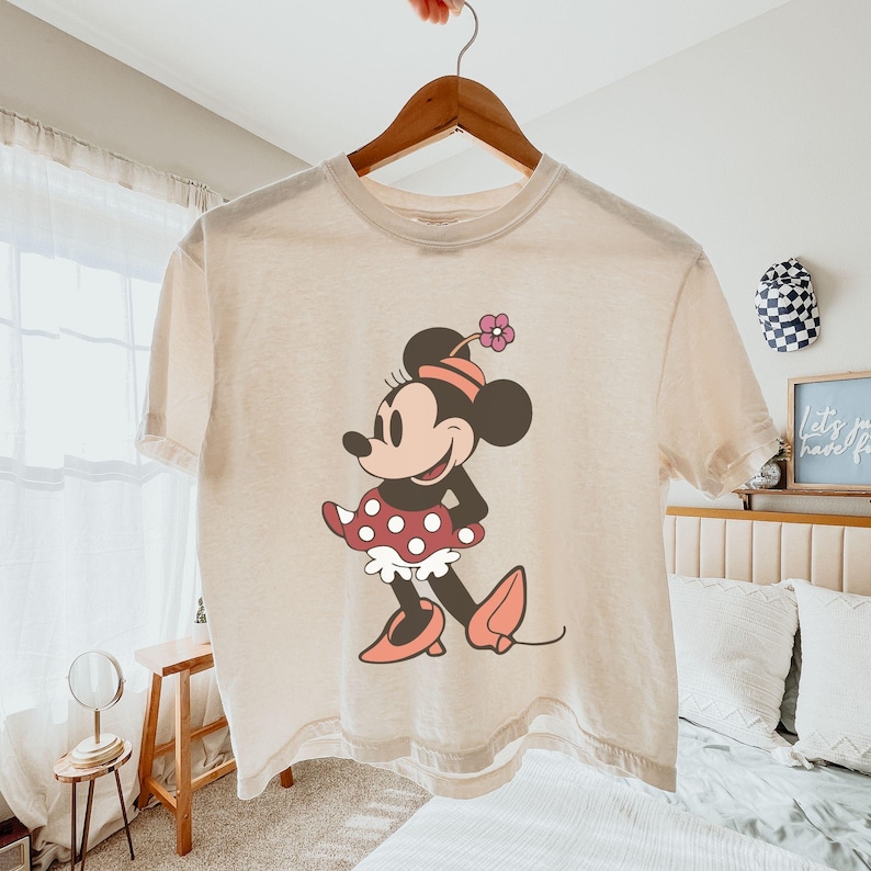 Op de afbeelding: Een beige t-shirt met een Minnie Mouse-afbeelding. Het stripfiguur wordt getoond in een klassieke pose, met een rode polkadot rok, roze schoenen en een bijpassende hoed met een bloem. Het t-shirt hangt aan een houten hanger.