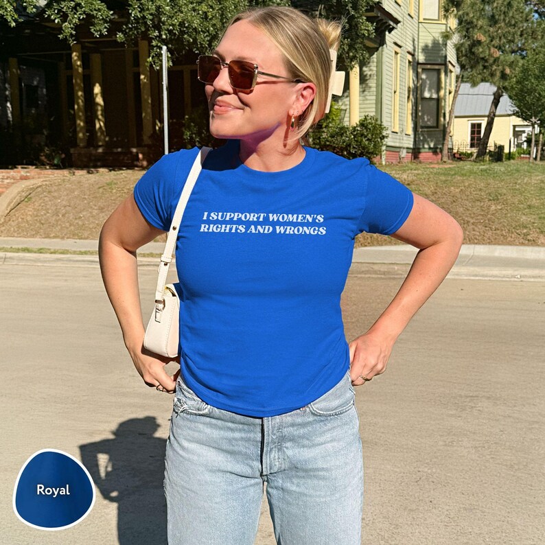 May include: Royal blue t-shirt with the text "I SUPPORT WOMEN'S RIGHTS AND WRONGS" in white. The shirt is paired with light blue jeans. A white purse is worn over the shoulder.