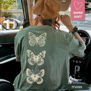 May include: A moss green t-shirt featuring three detailed butterfly illustrations in a vertical arrangement. The butterflies are in a cream color, contrasting against the shirt's background. The shirt is made of a soft material. The word "moss" is visible.