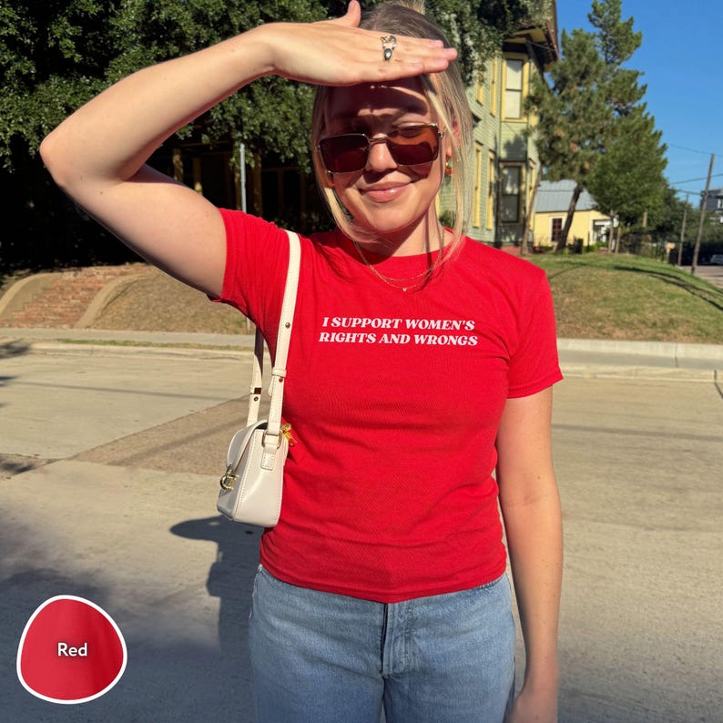 May include: A red t-shirt with the text "I SUPPORT WOMEN'S RIGHTS AND WRONGS" in white. The shirt is paired with blue jeans and a white purse with a shoulder strap. The image is taken outdoors on a sunny day.