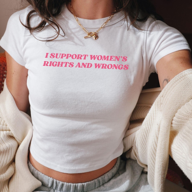 May include: White t-shirt with the pink text "I SUPPORT WOMEN'S RIGHTS AND WRONGS". The shirt has short sleeves and a crew neck. A gold necklace is visible. The shirt is paired with gray sweatpants and a cream-colored cardigan.