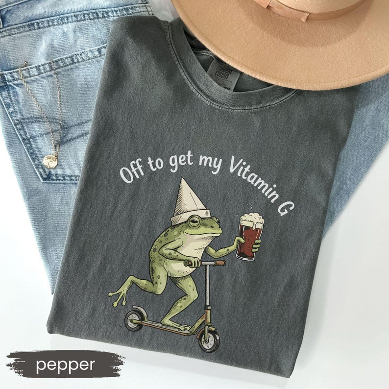 May include: Pepper-colored t-shirt with a graphic of a frog riding a scooter and holding a beer. The text "Off to get my Vitamin G" is printed above the frog. The shirt is laid on top of blue jeans and a tan hat.