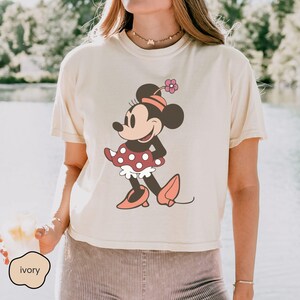 Op de afbeelding: Ivoor T-shirt met een Minnie Mouse-afbeelding. Het stripfiguur is afgebeeld in een rood-witte polkadotjurk, roze schoenen en een bijpassende hoed met een bloem. Het shirt heeft korte mouwen en een relaxte pasvorm.