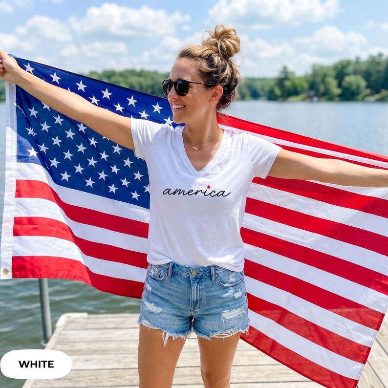 K&ouml;nnte beinhalten: Ein wei&szlig;es V-Ausschnitt-T-Shirt mit dem Wort "america" in Schreibschrift, kombiniert mit Jeansshorts. Die Person h&auml;lt eine gro&szlig;e amerikanische Flagge mit roten und wei&szlig;en Streifen und blauen Sternen.