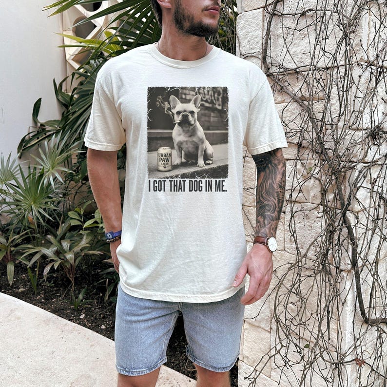 K&ouml;nnte beinhalten: Cremefarbenes T-Shirt mit einem Schwarz-Wei&szlig;-Bild einer Franz&ouml;sischen Bulldogge und einer Dose "Urban Paw" Bier. Der Text "I GOT THAT DOG IN ME" ist darunter gedruckt. Das Shirt wird mit blauen Jeansshorts kombiniert.