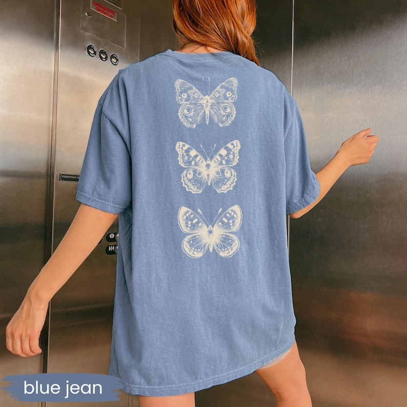 May include: A blue jean colored t-shirt with three cream-colored butterfly illustrations on the back. The t-shirt is a relaxed fit and has short sleeves. The text "blue jean" is at the bottom of the image.
