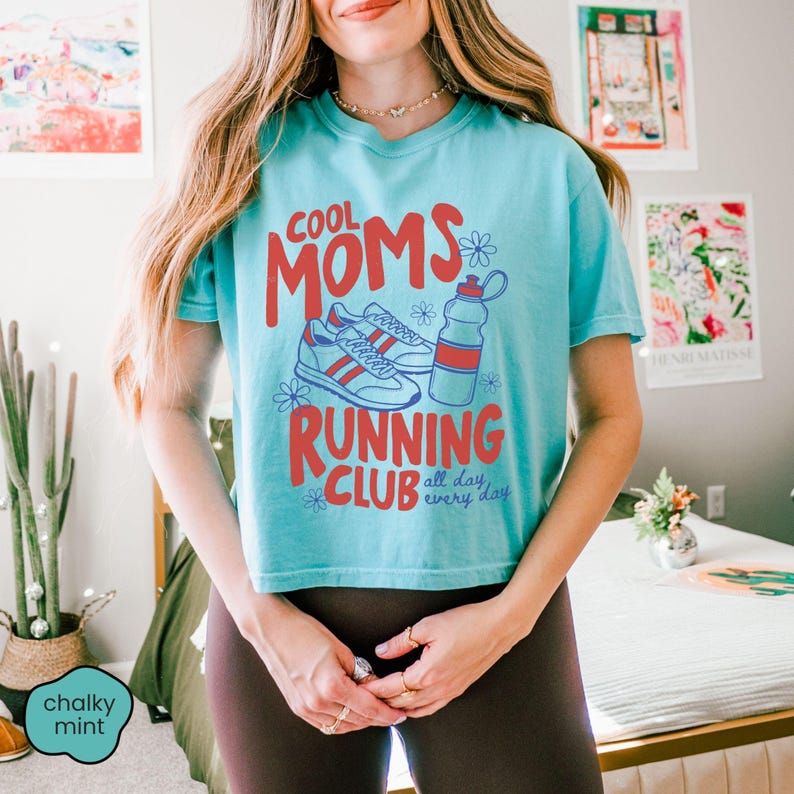 May include: A teal short-sleeved t-shirt featuring red text that reads COOL MOMS RUNNING CLUB all day every day. The graphic includes a pair of running shoes, a water bottle, and small blue flower outlines centered on the front of the garment.