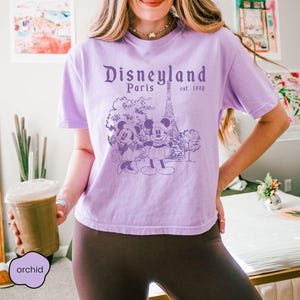 Puede incluir: Camiseta de color orqu&iacute;dea con un dise&ntilde;o de Disneyland Par&iacute;s que incluye a Mickey y Minnie Mouse, la Torre Eiffel y el texto "est. 1992". La camiseta tiene mangas cortas y cuello redondo.