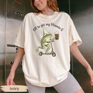 May include: Ivory-colored t-shirt featuring a whimsical graphic of a frog riding a scooter while holding a beer. The text "Off to get my Vitamin G" is printed above the frog. The shirt has a relaxed fit.