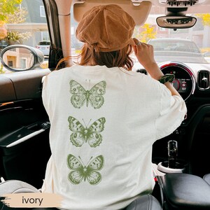 May include: Ivory-colored t-shirt featuring three green butterfly illustrations on the back. The shirt is a relaxed fit. The word "ivory" is printed at the bottom of the image.