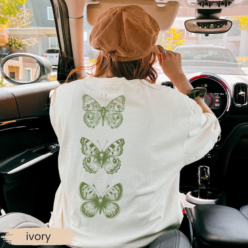 May include: Ivory-colored t-shirt featuring three green butterfly illustrations on the back. The shirt is a relaxed fit. The word "ivory" is printed at the bottom of the image.