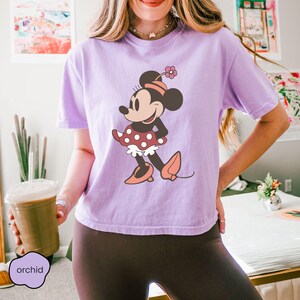 Op de afbeelding: Een lila T-shirt met een Minnie Mouse-afbeelding. Het stripfiguur draagt een rode polkadotjurk, bijpassende schoenen en een met bloemen versierde hoofdband. Het woord "orchid" is gedrukt op een klein, lila label.