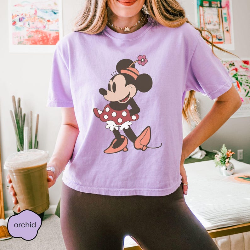 Op de afbeelding: Een lila T-shirt met een Minnie Mouse-afbeelding. Het stripfiguur draagt een rode polkadotjurk, bijpassende schoenen en een met bloemen versierde hoofdband. Het woord "orchid" is gedrukt op een klein, lila label.