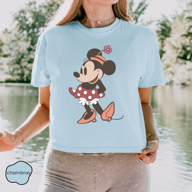 Op de afbeelding: Lichtblauw T-shirt met een cartoon illustratie van Minnie Mouse. Minnie draagt een rode polkadot jurk met een witte ruche, roze schoenen en een bloem op haar hoofd. Het shirt heeft een ronde hals en korte mouwen.