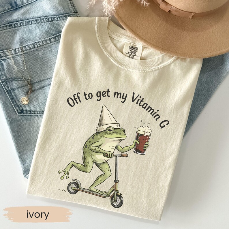 May include: Ivory-colored t-shirt with the text "Off to get my Vitamin G." The graphic shows a green frog wearing a chef's hat, riding a scooter, and holding a glass of beer. The shirt is folded on top of blue jeans.